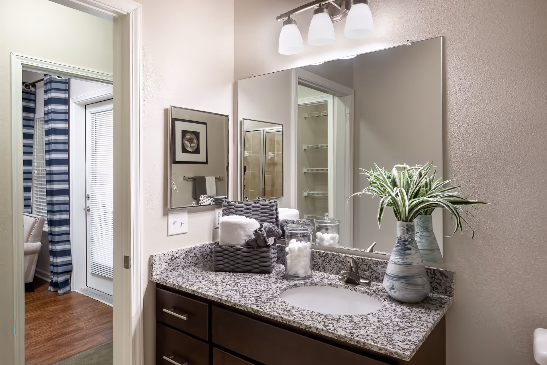 Bathroom vanity with a granite countertop, an oval sink, and a silver faucet. On the countertop are a blue and white vase with green and white leaves, two glass jars filled with cotton balls, and a woven basket holding rolled towels. Above the vanity is a large mirror with three light fixtures. The bathroom door is open, showing a glimpse of a room with wooden flooring, a white door with blinds, and blue and white striped curtains.