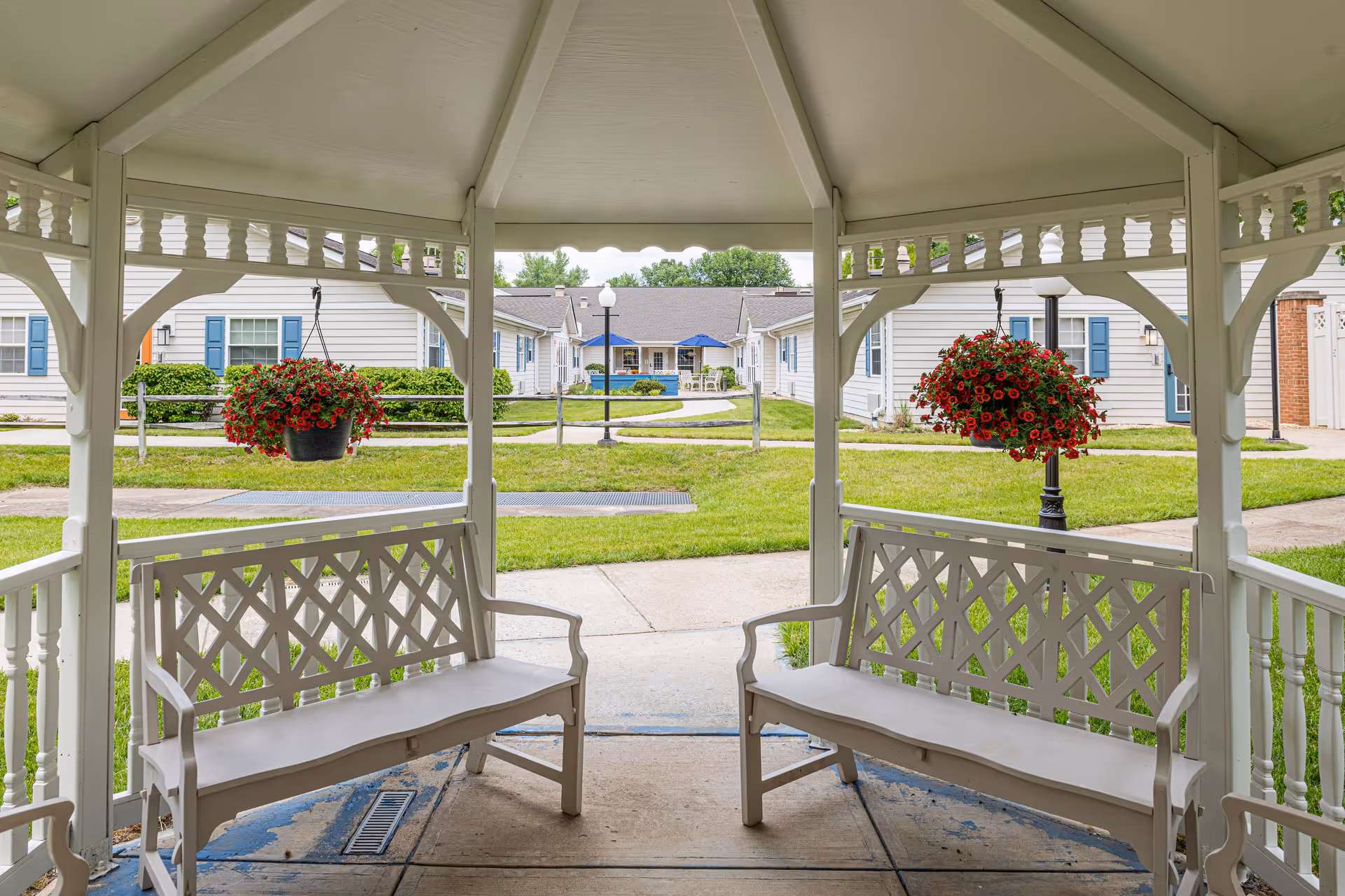 View from inside a white gazebo with two white benches facing each other and hanging baskets of red flowers. Outside the gazebo, there is a paved walkway, green grass, and several single-story buildings with white siding and blue shutters.