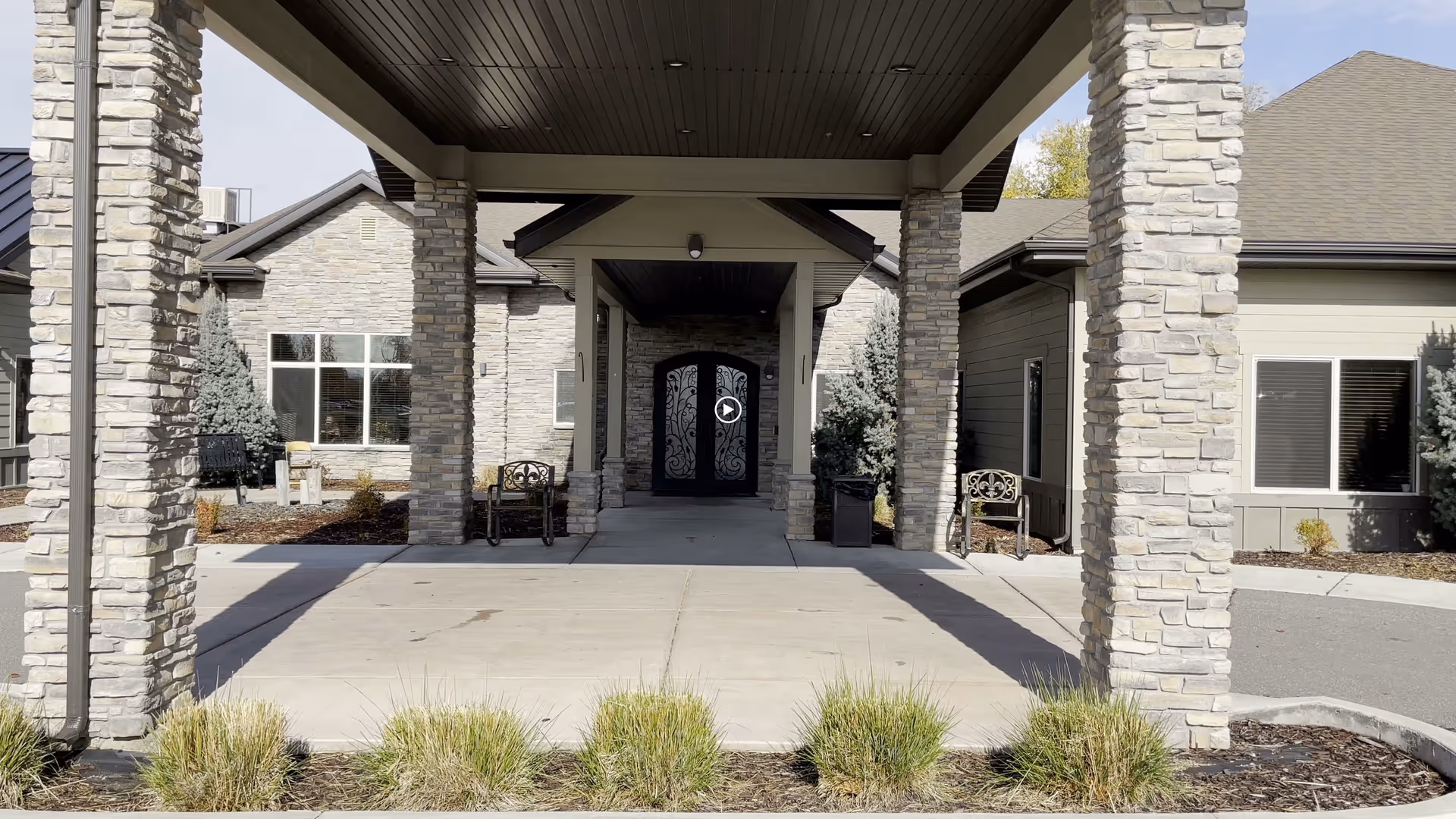 Covered entrance of a stone-faced assisted living building with pillars, benches, and decorative double doors.