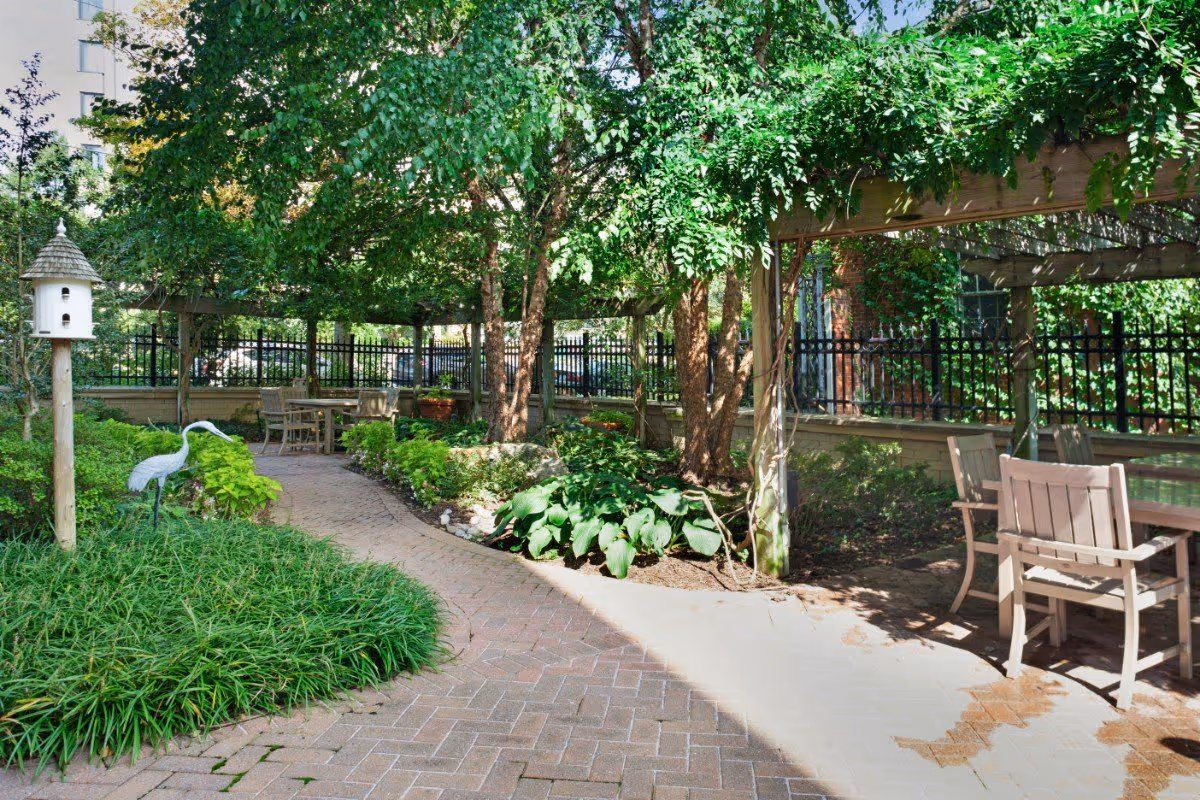 Shaded outdoor courtyard with a brick walkway, pergola-covered seating areas, lush plantings and a decorative birdhouse.