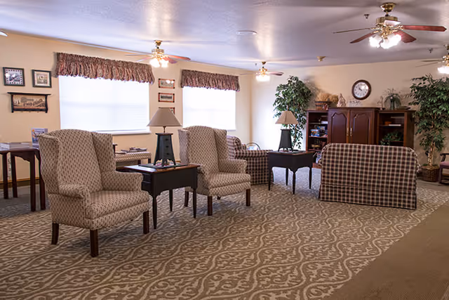 Well-lit senior living common room with upholstered armchairs, side tables, sofas, lamps, and a TV cabinet.