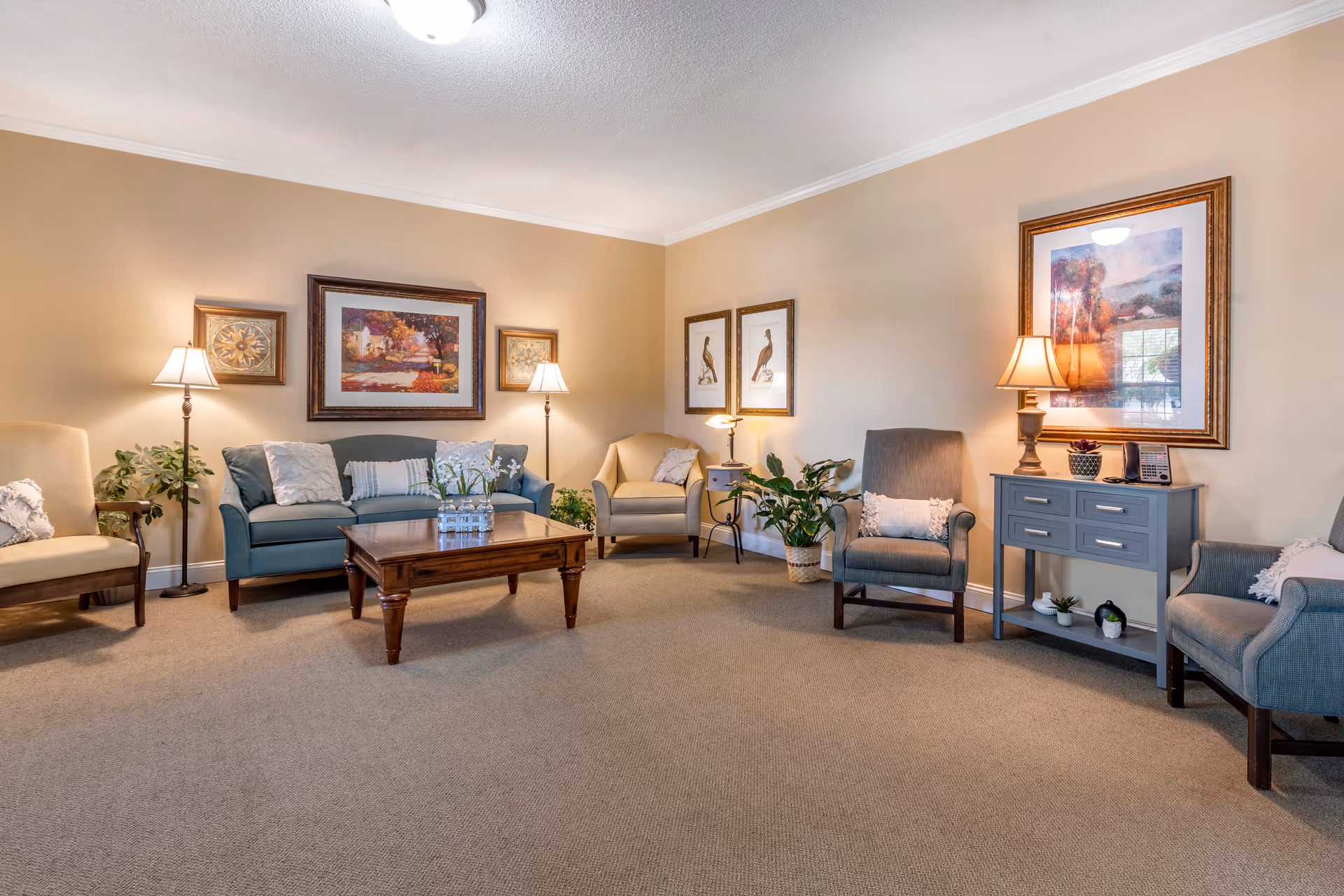A cozy living room with beige walls and carpet, featuring a blue sofa, several armchairs, a wooden coffee table with a glass vase of flowers, floor lamps, framed artwork on the walls, and a small gray side table with a lamp and decorative items.