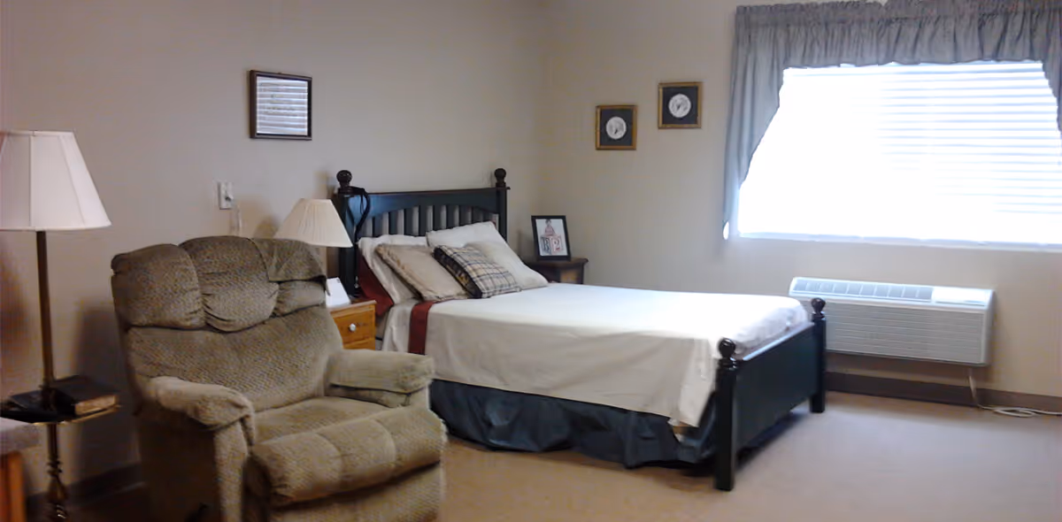A bedroom in a senior living facility with a single bed made up with white sheets and multiple pillows. Next to the bed is a wooden nightstand with a lamp and a framed photo. A comfortable recliner chair is positioned near the bed, and a floor lamp stands beside it. A large window with blinds and a curtain allows natural light into the room. The walls are decorated with a few small framed pictures.