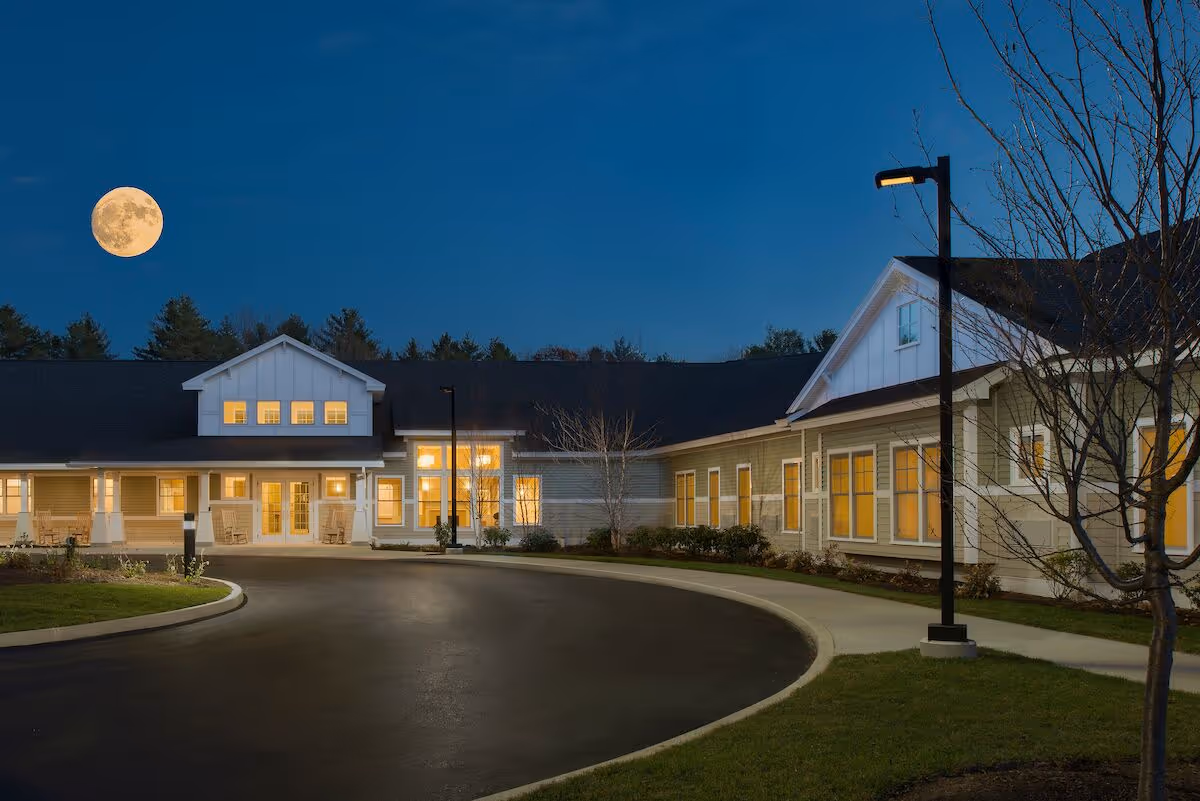 Exterior view of Avita of Wells senior living facility at night with lights glowing from windows, a curved driveway, street lamps, and a full moon in the clear sky.