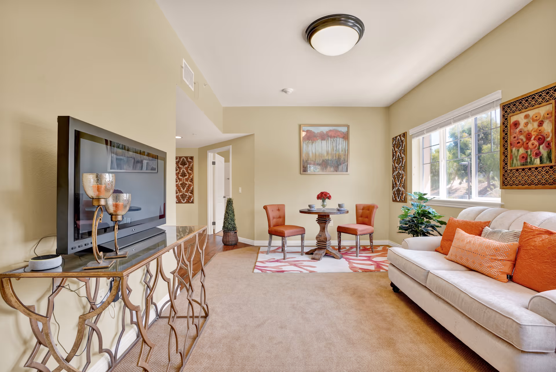 A cozy living room with a beige sofa adorned with orange and patterned pillows, a small round wooden table with two orange chairs on a patterned rug, a flat-screen TV on a decorative metal console table, and wall art including floral and tree paintings. A large window lets in natural light, and the walls are painted a soft beige.