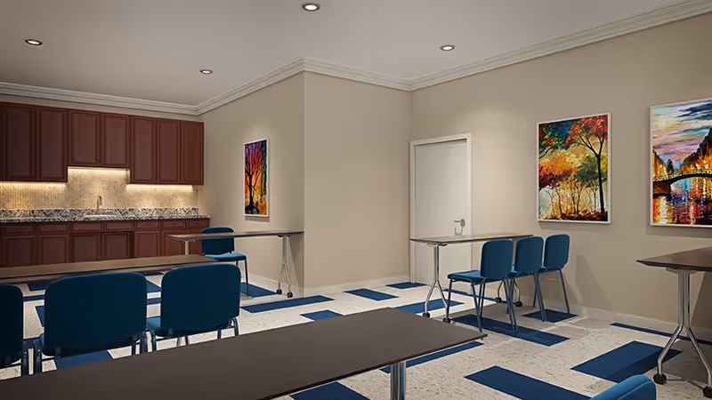 Interior view of a room with multiple tables and blue chairs arranged in rows. The room has beige walls adorned with colorful paintings and a kitchenette area with wooden cabinets and a granite countertop along one wall. The floor has a patterned design with blue and white tiles.