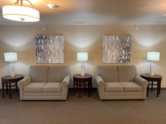 Two beige loveseats separated by a round wooden side table with a lamp on it, each loveseat flanked by a similar side table and lamp. Above each loveseat hangs a large abstract painting with neutral tones. The room has beige walls and carpeted floor, with a ceiling light fixture visible.