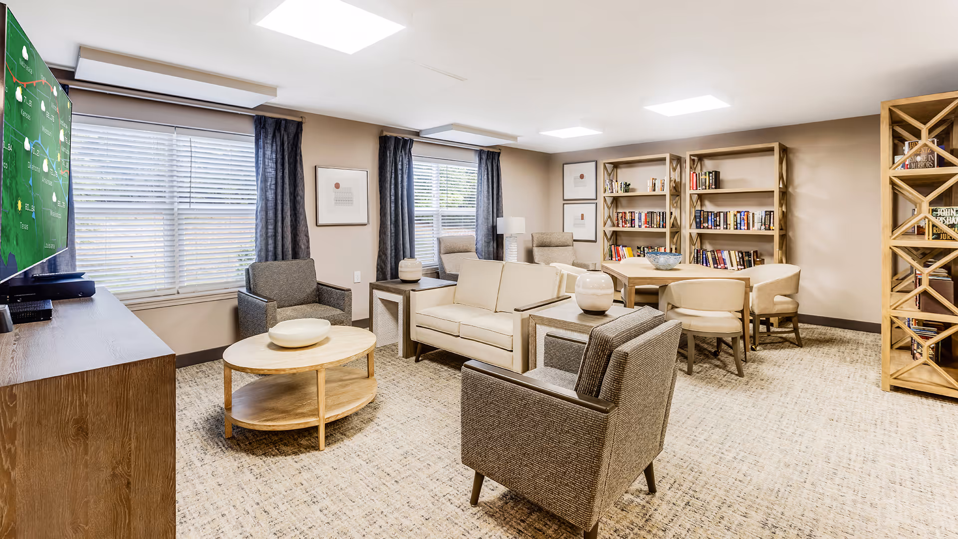 A bright and cozy living room area in a senior living facility featuring a beige sofa, two gray armchairs, a round wooden coffee table, and a wooden TV stand with a large flat-screen TV. The room has large windows with blinds and dark curtains, beige walls, carpeted floor, and wooden bookshelves filled with books and decorative items. There is also a small round table with four chairs near the bookshelves.