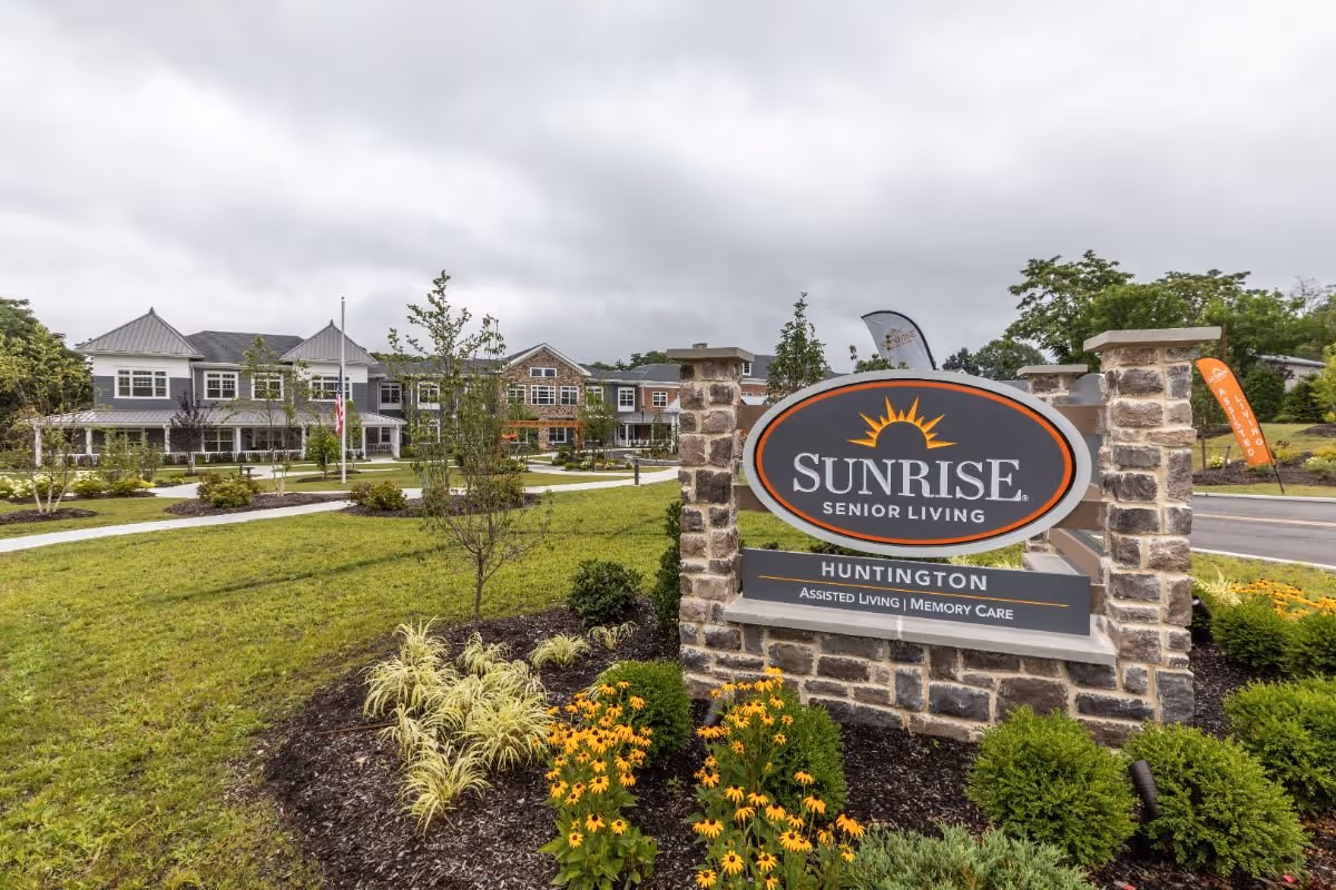 Outdoor view of Sunrise Senior Living facility in Huntington, showing a large stone sign with the facility name and description, surrounded by landscaped greenery and flowers, with the building and cloudy sky in the background.