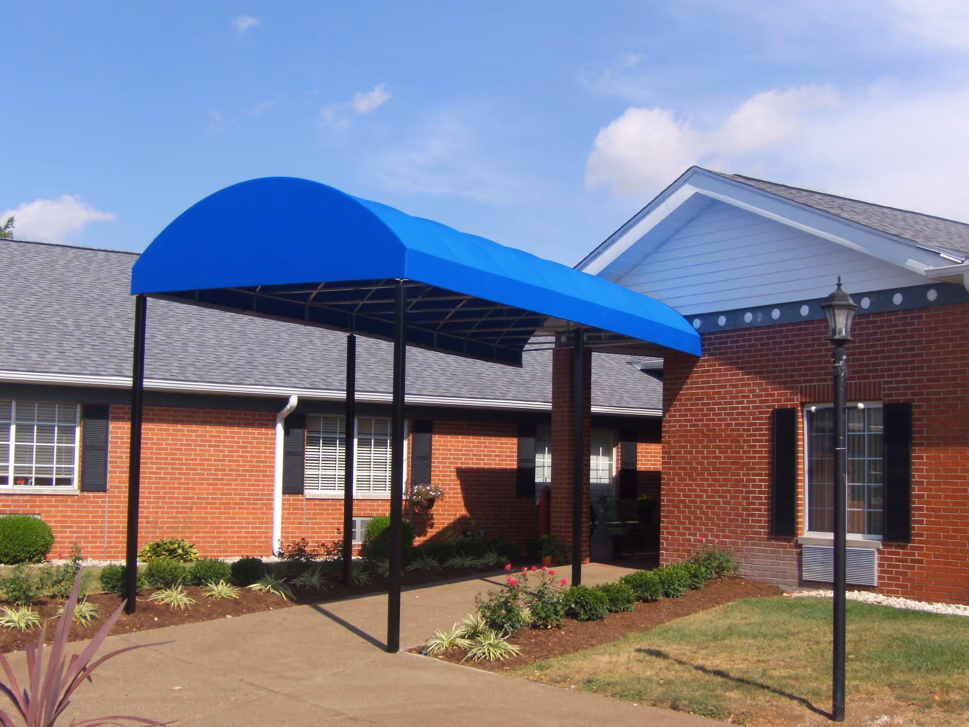 Entrance to a brick building with a blue canopy over the walkway, surrounded by landscaping with bushes and flowers, under a partly cloudy sky.