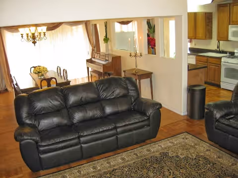 Interior view of a living room area with a black leather sofa in the foreground, a patterned rug on the wooden floor, and a dining area with a table and chairs in the background. There is a piano against the wall, a chandelier hanging above the dining table, and a kitchen area with wooden cabinets and white appliances visible to the right.