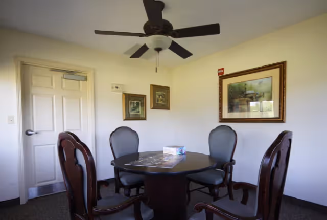 Small dining/meeting room with a round wooden table surrounded by four upholstered chairs, a ceiling fan, framed artwork on the walls, and a closed door.