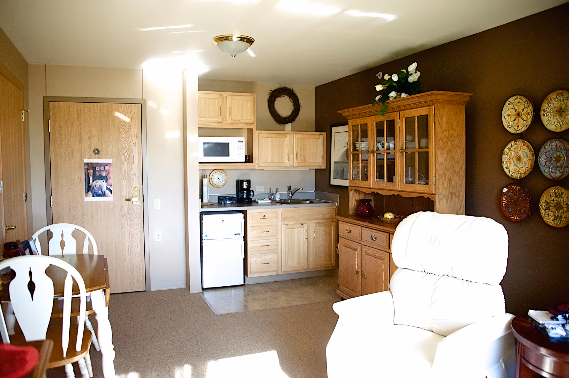 A cozy interior room featuring a small kitchenette with light wood cabinets, a microwave, mini fridge, and coffee maker. To the right, there is a wooden hutch with glass doors displaying dishes and decorative items. A comfortable white recliner chair is positioned in the foreground. On the left side, a wooden dining table with white chairs is partially visible. The walls are painted brown and beige, adorned with decorative plates and a wreath. A closed wooden door is seen in the background.