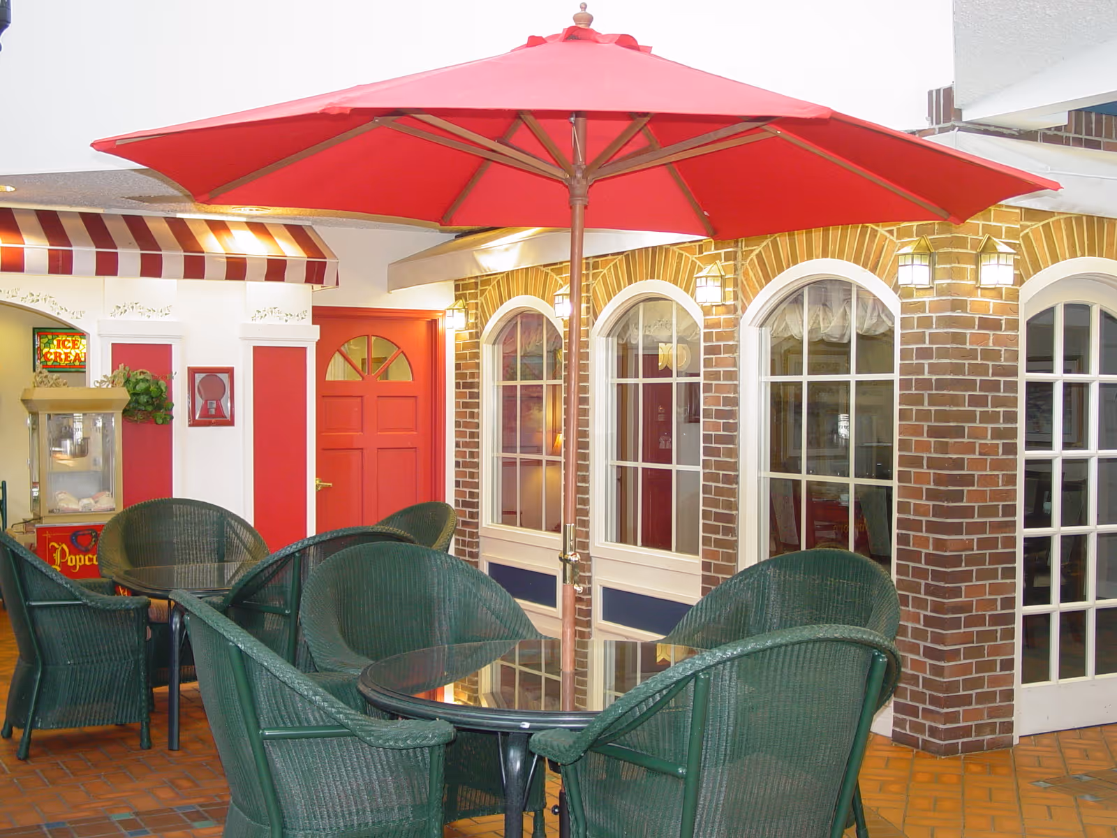 Indoor seating area with a round glass table surrounded by green wicker chairs and a large red umbrella overhead. The background features a red door, brick walls with arched windows, and a popcorn machine under a red and white striped awning.