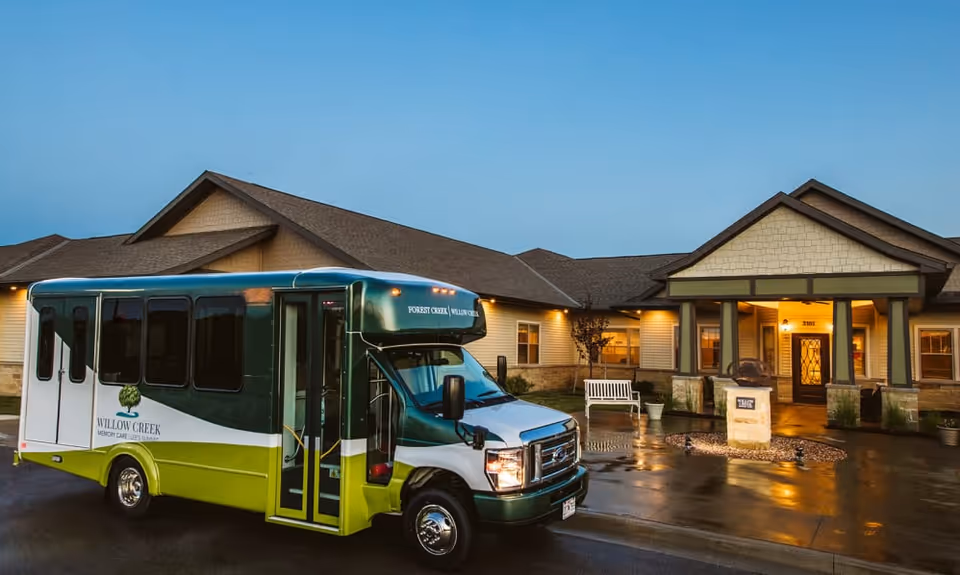 A green and white shuttle bus parked in front of a single-story senior living facility building at dusk. The building has a welcoming entrance with a covered porch, stone accents, and warm lighting. The pavement is wet, reflecting the lights from the building and bus.