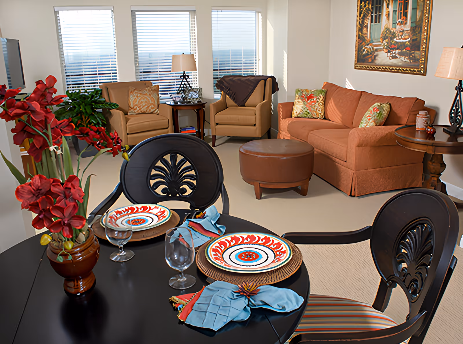 A cozy living room and dining area with a round black dining table set for two with colorful plates, glasses, and blue napkins. The living room has a brown sofa with patterned cushions, two beige armchairs, a round brown ottoman, side tables with lamps, a large painting on the wall, and large windows with blinds letting in natural light. A vase with red flowers is on the dining table.