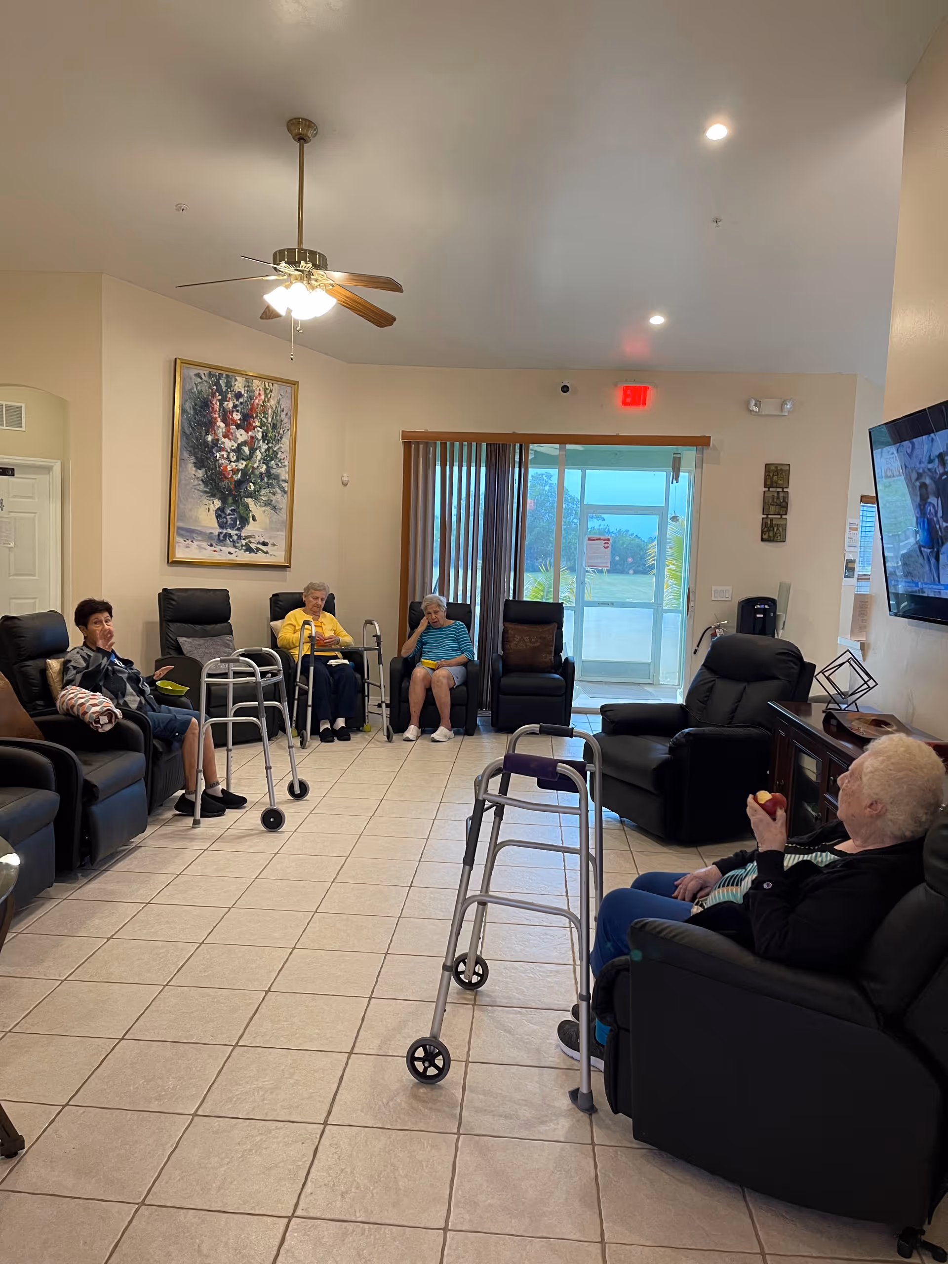 A group of elderly people sitting in a spacious, well-lit common room with tiled floors and beige walls. They are seated in comfortable black recliners arranged in a semi-circle, some with walkers nearby. A ceiling fan with lights hangs from the ceiling, and a large painting of flowers is on the wall. A flat-screen TV is mounted on the right wall, and a glass door with vertical blinds leads outside.