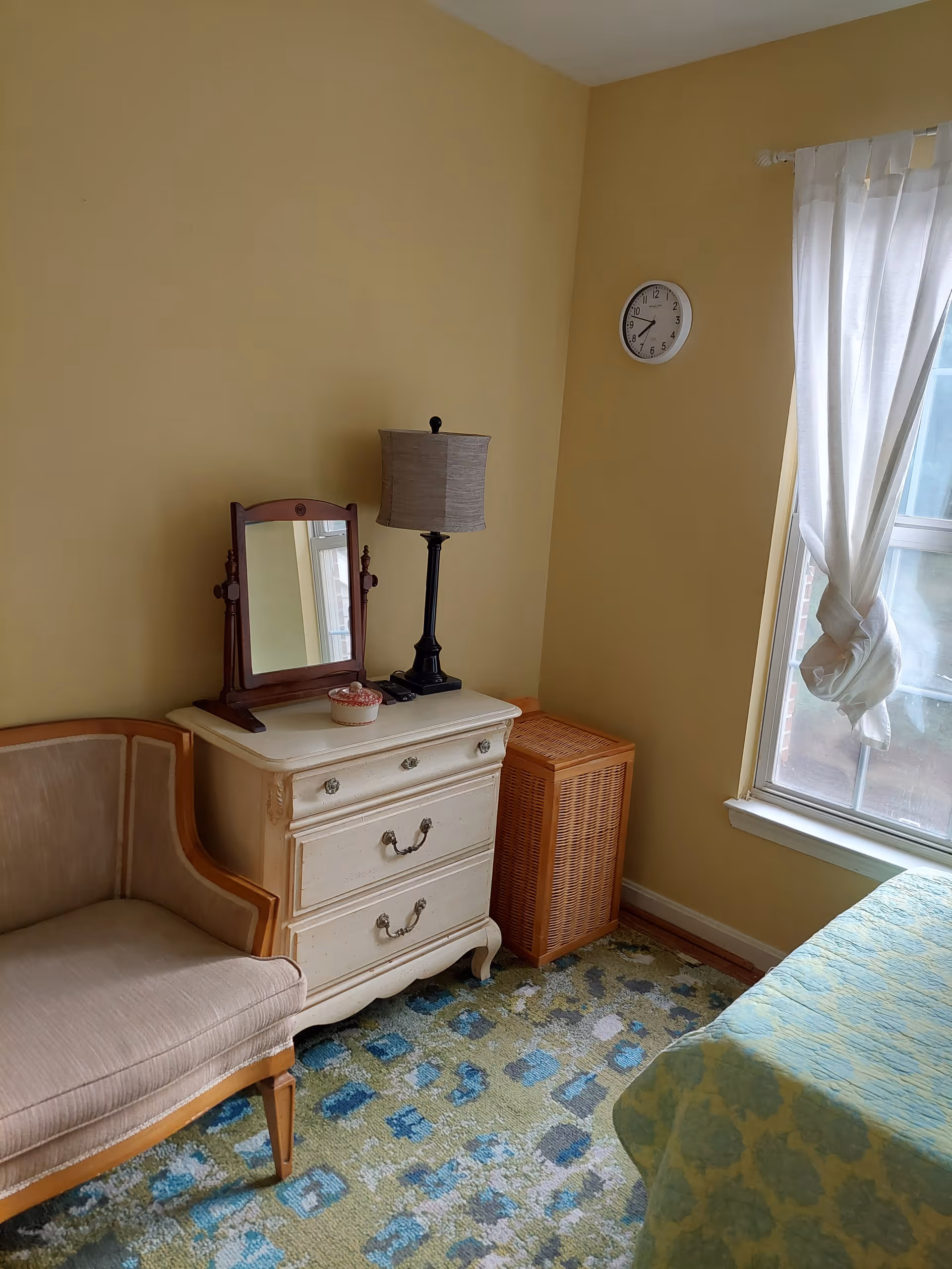 A cozy corner of a bedroom with yellow walls, featuring a beige upholstered chair, a white wooden dresser with three drawers, a table lamp with a beige shade, a small mirror, a wicker laundry basket, a window with white curtains tied in a knot, and part of a bed with a green and yellow patterned bedspread. A round wall clock shows the time as 8:35.