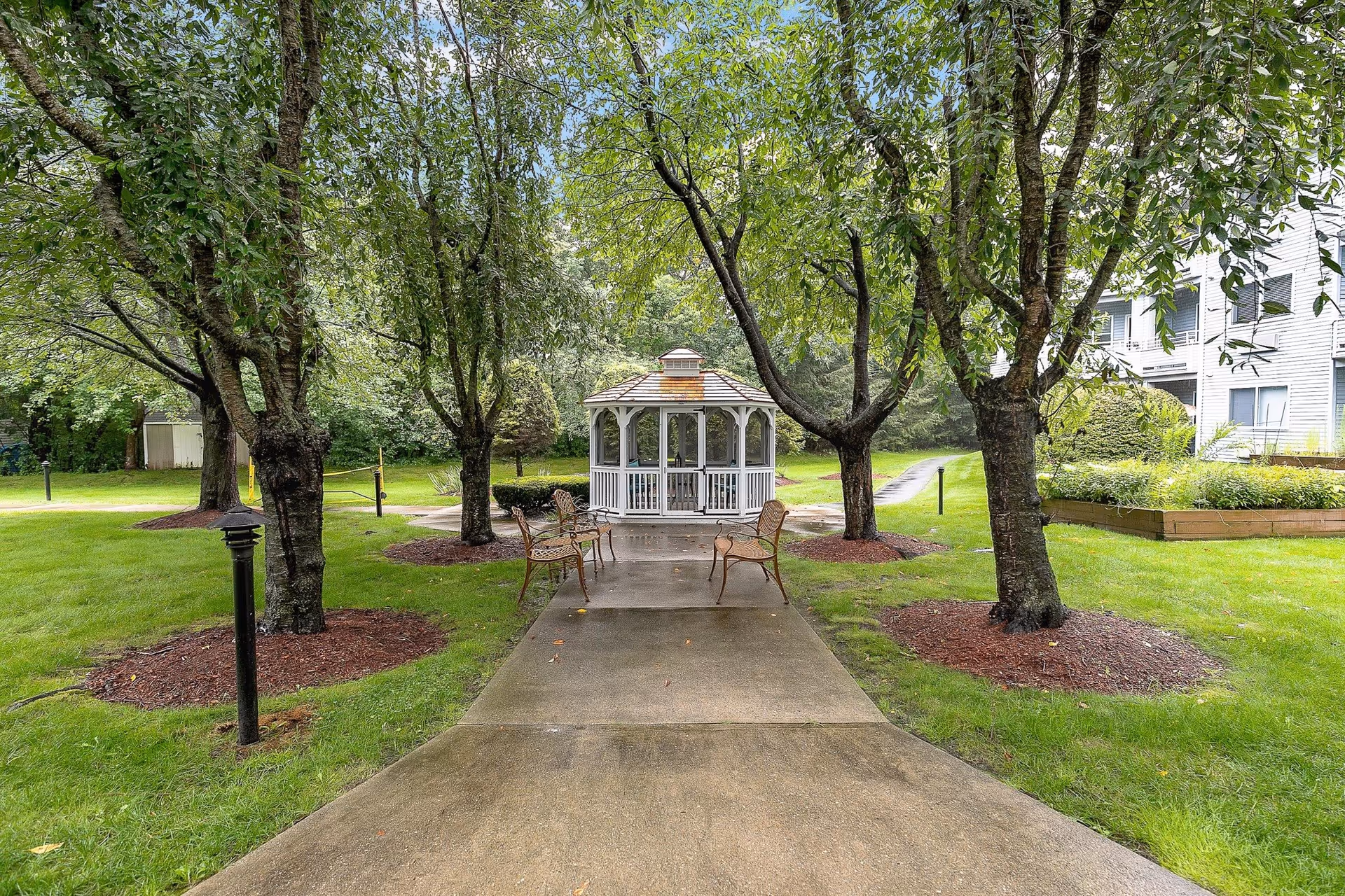 A paved walkway lined with trees leads to a white gazebo with a shingled roof in a green outdoor garden area. There are metal chairs placed along the walkway near the gazebo. The surrounding area is grassy with mulched tree bases and a building is partially visible on the right side.