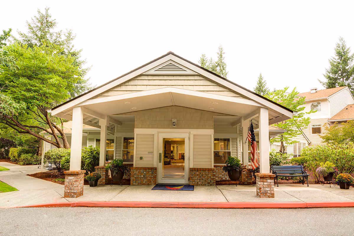 Front entrance of a senior living building with a covered portico, brick columns, potted plants, and an American flag.