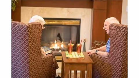 Two people sit in patterned armchairs facing each other by a lit fireplace with a small table holding two drinks.