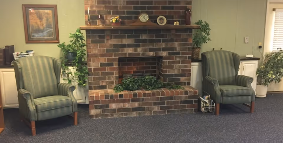A cozy interior room featuring a brick fireplace with a wooden mantel decorated with a clock, small framed picture, and a vase. Two green upholstered armchairs are positioned on either side of the fireplace. There are potted plants and framed artwork on the walls, with cabinets and a window with blinds in the background.