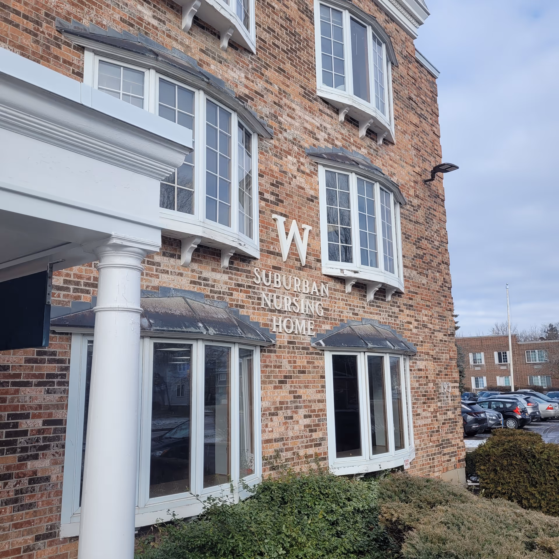 Exterior view of a brick building with white-framed windows and a white column. The building has a sign that reads 'W Suburban Nursing Home'. There are bushes in front of the building and a parking lot with cars in the background under a cloudy sky.