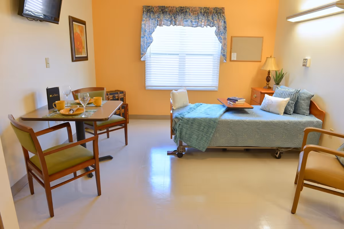 A bright room with a single bed covered in a blue patterned bedspread and several pillows. Next to the bed is a wooden nightstand with a lamp and a plant. A small table with two chairs is set with cups, glasses, and a menu. A window with blue floral curtains lets in natural light. The walls are painted a warm yellow color, and a flat-screen TV is mounted on the wall above a framed picture.