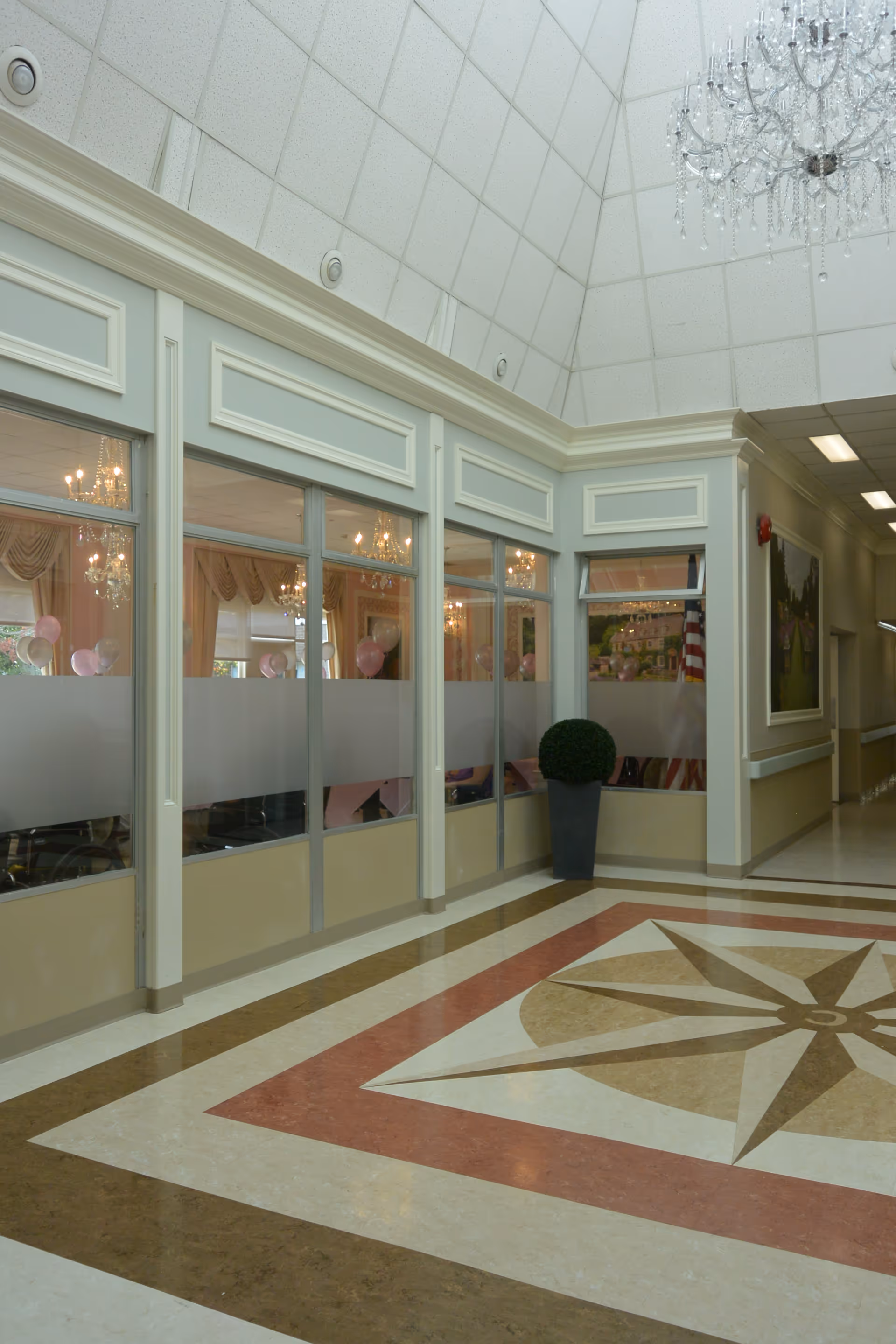 Interior hallway of a nursing and rehabilitation center with a decorative patterned floor, large windows with frosted glass panels, a potted plant, chandeliers visible through the windows, and a high ceiling with a chandelier.