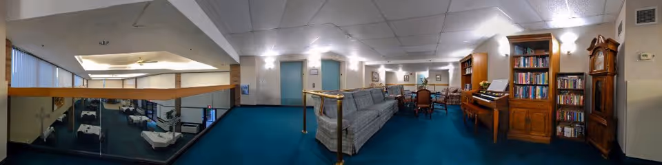Panoramic view of a senior living common lounge with couches, bookshelves, a piano, seating tables and a railing overlooking a lower dining area.