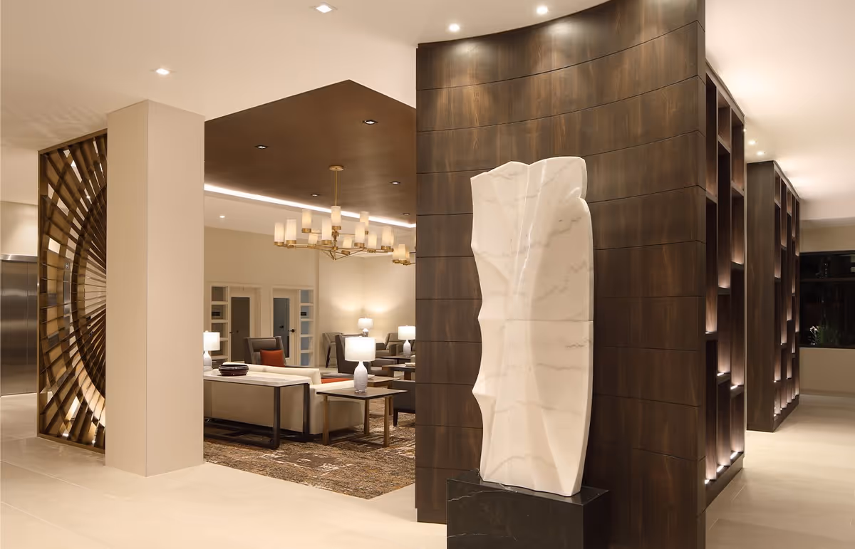 A modern interior space featuring a large abstract white sculpture on a black pedestal in front of a dark wood-paneled wall. To the left, there is a seating area with beige sofas, side tables with lamps, and a chandelier hanging from a dark ceiling section. The floor is light-colored tile, and there is a decorative wooden partition near an elevator.