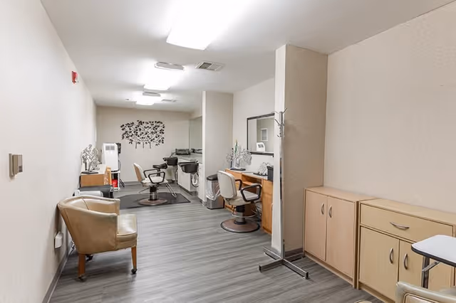 Interior view of a hair salon area in a senior living facility with salon chairs, mirrors, hair washing stations, a coat rack, and cabinets along the walls. The floor is covered with wood-like vinyl flooring and the walls are painted light beige. There is a decorative tree wall decal at the far end of the room.