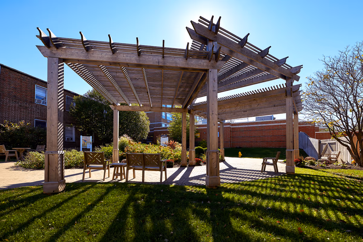 Outdoor seating area with wooden pergolas casting shadows on the grass and paved ground, surrounded by greenery and brick buildings under a clear blue sky.