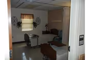 A small, simple room with two hospital-style beds, a window with a valance curtain, a small table with a chair, and wall decorations including two circular metal art pieces and some greenery. The room number 609 is visible on the door frame.