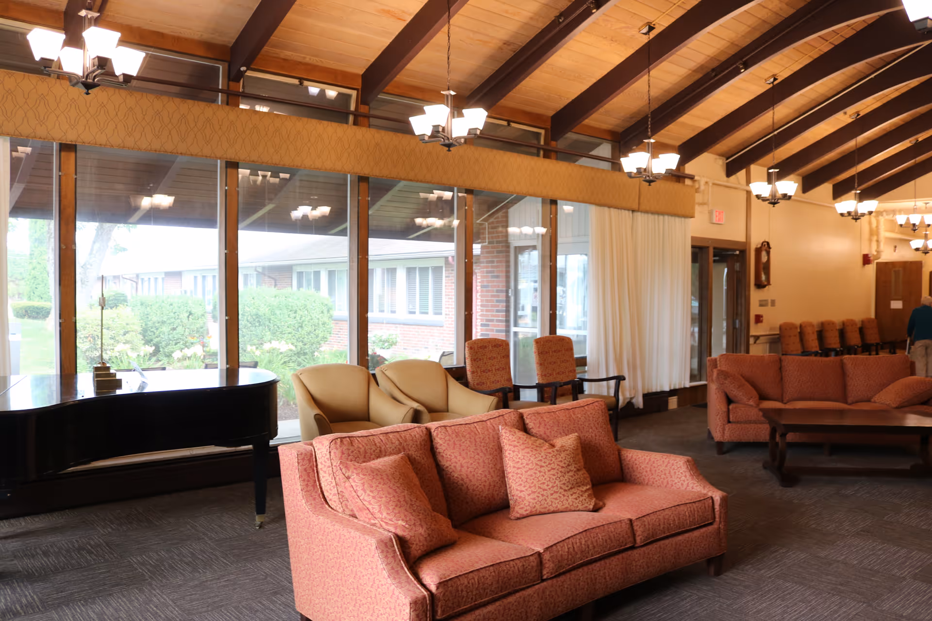 A spacious living room area with large windows showing an outdoor view. The room features two coral-colored sofas with matching cushions, two beige armchairs, and two patterned chairs. A black grand piano is positioned near the windows. The ceiling has exposed wooden beams and multiple hanging light fixtures. There is a wooden coffee table in front of one of the sofas and a row of chairs lined up against the wall in the background.