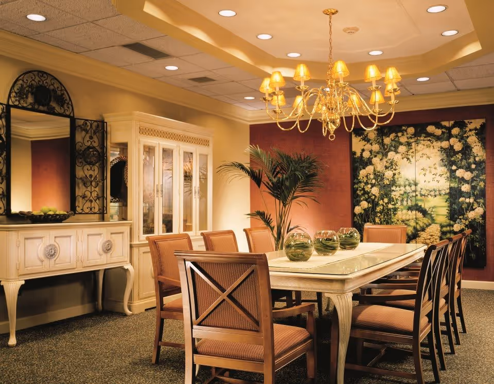 A warmly lit dining room with a long rectangular table surrounded by eight cushioned wooden chairs. On the table are three glass bowls with green plants inside. A large chandelier with multiple lampshades hangs above the table. In the background, there is a decorative cabinet with glass doors, a large mirror with an ornate black frame, a potted plant, and a large floral painting on the wall.