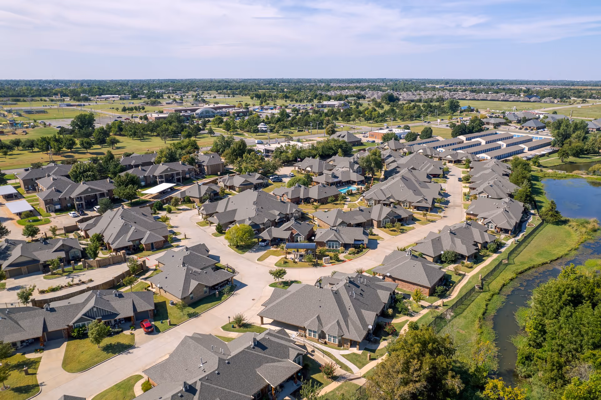 Aerial view of a residential community with multiple single-story houses arranged along curved streets, surrounded by green lawns, trees, and a small pond on the right side. The background shows more buildings, open fields, and a partly cloudy sky.