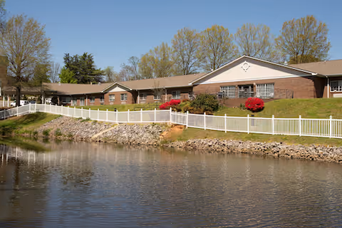Exterior view of a single-story brick building with a white fence along a rocky shoreline beside a calm body of water. Trees with early spring foliage are visible behind the building under a clear blue sky.