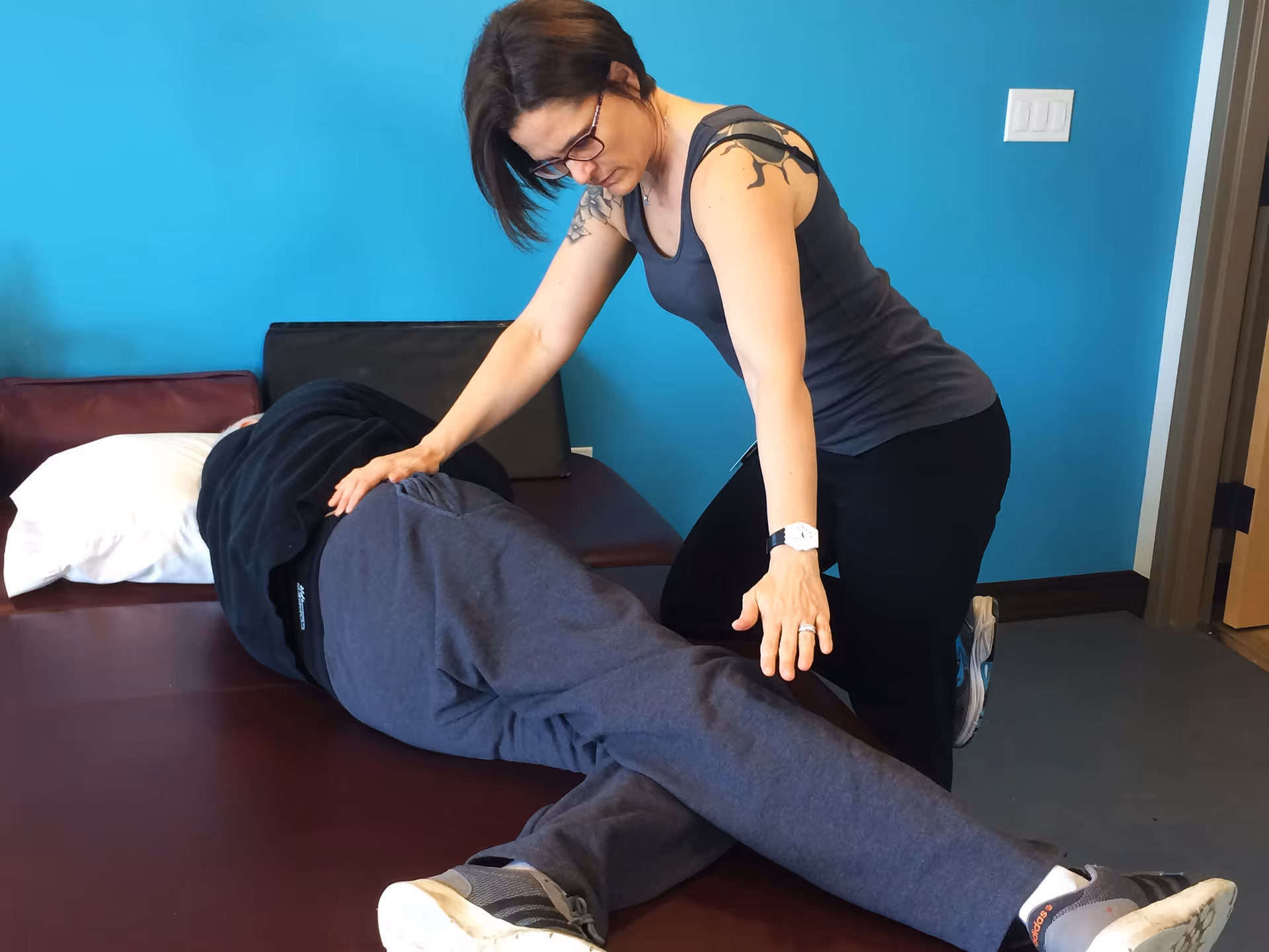 A woman wearing glasses and a sleeveless top assists a person lying on their side on a padded table, possibly helping with a physical therapy or stretching exercise in a room with a blue wall.