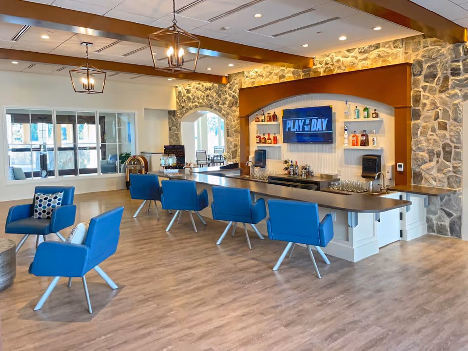A modern lounge area in a senior living facility featuring a bar with blue cushioned chairs, a stone accent wall, and a TV screen displaying 'PLAY OF THE DAY'. The room has wooden flooring, pendant lights, and a windowed wall looking into another seating area.