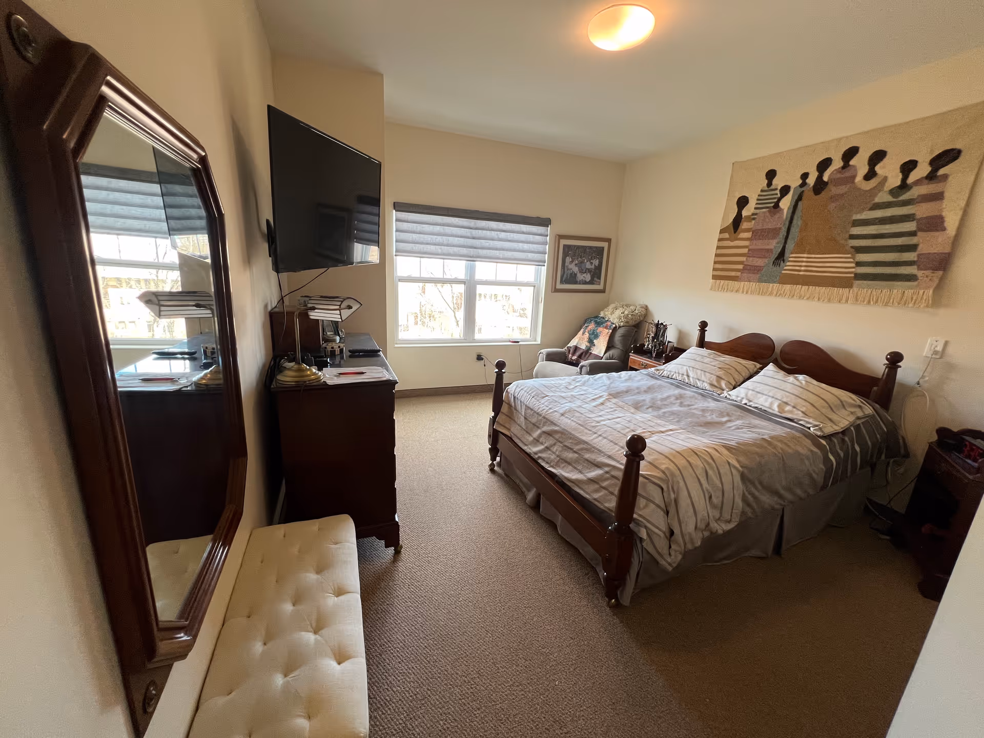 A bedroom with a wooden bed frame and striped bedding, a cushioned bench, a wall-mounted flat screen TV, a dresser with a lamp, a comfortable armchair with a blanket, a framed picture on the wall, and a decorative wall hanging featuring stylized human figures. The room has beige walls and carpeted floor with a window letting in natural light.