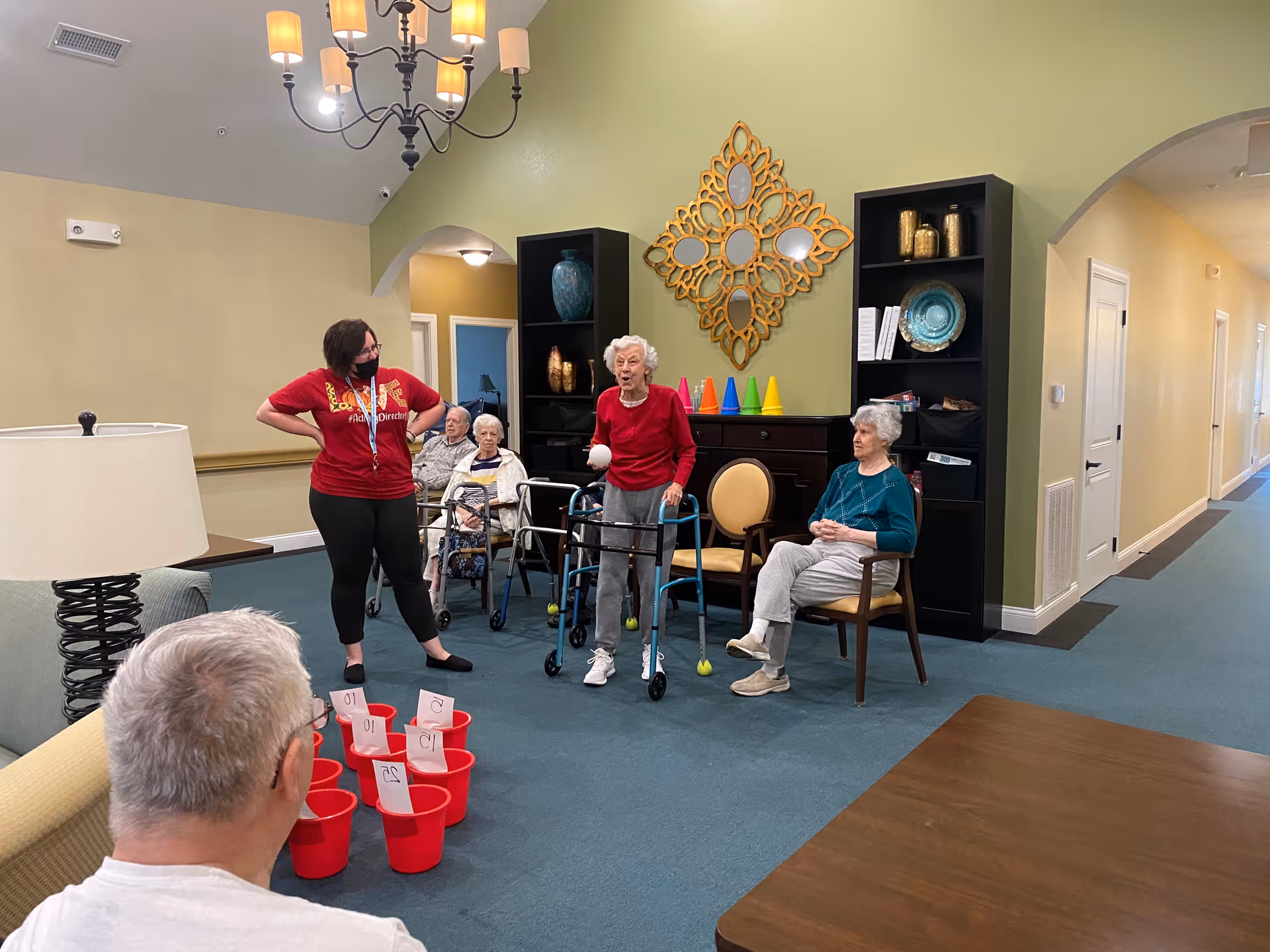 A group of elderly residents and a staff member in a common area of a senior living facility. The staff member, wearing a red shirt and black pants, stands with hands on hips. One elderly woman with a walker is holding a ball, appearing to participate in a game involving red buckets with numbers on them. Other residents are seated and watching. The room has green walls, blue carpet, a chandelier, and decorative shelves with vases and a mirror.