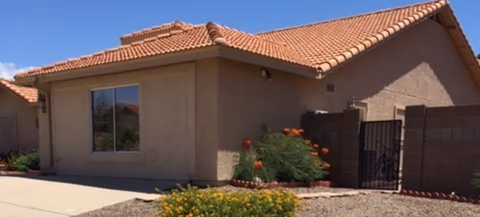 Exterior view of a single-story building with beige stucco walls and a red tile roof under a clear blue sky. There is a window on the visible wall and a gated entrance to a small enclosed area with some plants and flowers in front.