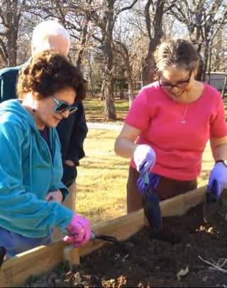 Three elderly people gardening outdoors in a raised garden bed. Two women in the foreground are wearing gloves and using small gardening tools to work the soil, while a man stands behind them. Trees and grass are visible in the background.