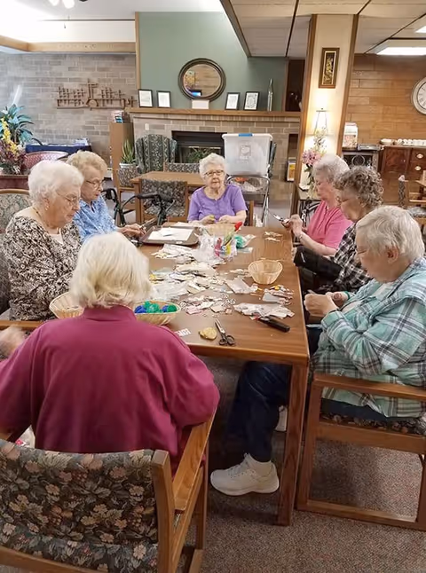 A group of elderly women sitting around a rectangular wooden table in a cozy room, engaging in a craft activity with various materials spread out on the table. The room has a fireplace, decorative items, and comfortable chairs, creating a warm and inviting atmosphere.