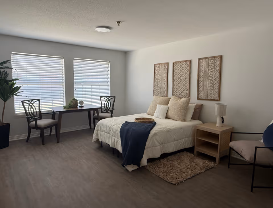 Bright, minimally decorated bedroom with a bed, nightstand, desk and two chairs by large windows.