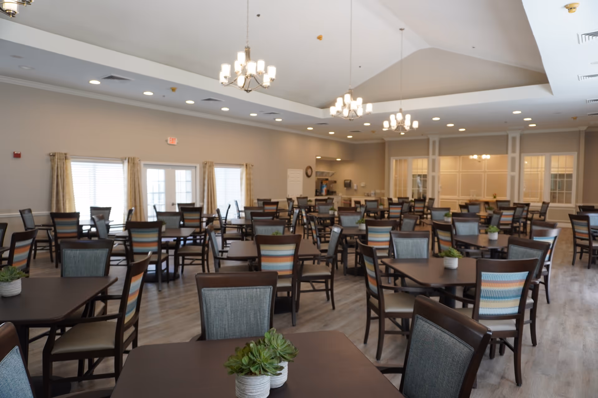 A spacious dining room with multiple dark wood tables and chairs featuring striped and solid upholstery. The room has large windows with curtains, chandeliers hanging from a tray ceiling, and small potted plants on the tables. The floor is light wood, and the walls are painted a soft neutral color.