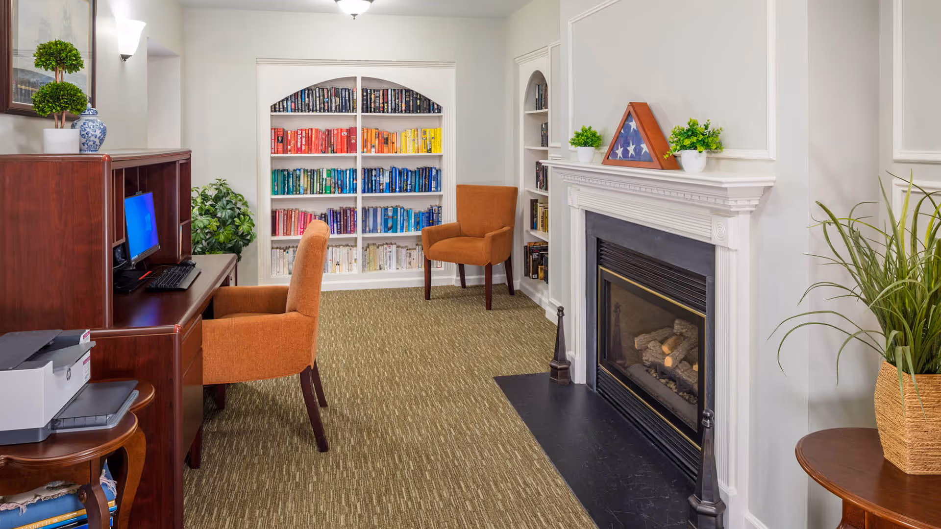 A cozy interior room featuring a fireplace with a white mantel decorated with a folded American flag and small potted plants. To the left, there is a wooden desk with a computer and an orange upholstered chair. Behind the desk, a built-in bookshelf is filled with books arranged by color in a rainbow pattern. The room has beige carpet and white walls with decorative molding.