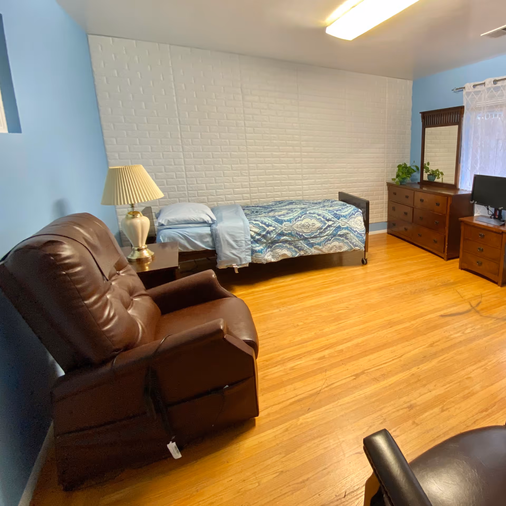 A bedroom with light blue walls and wooden floor featuring a single bed with blue patterned bedding, a wooden dresser with a mirror, a small TV on a wooden stand, a brown leather recliner chair, and a table lamp on a bedside table.