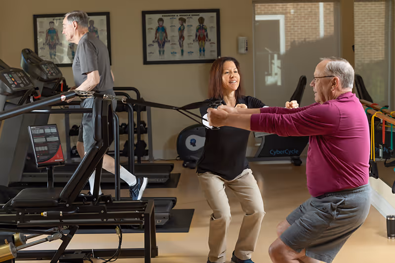 An elderly man in a magenta shirt and gray shorts is exercising with resistance bands assisted by a female trainer in a black shirt and beige pants in a fitness room. Another elderly man is walking on a treadmill in the background. Anatomical charts are visible on the wall behind them.
